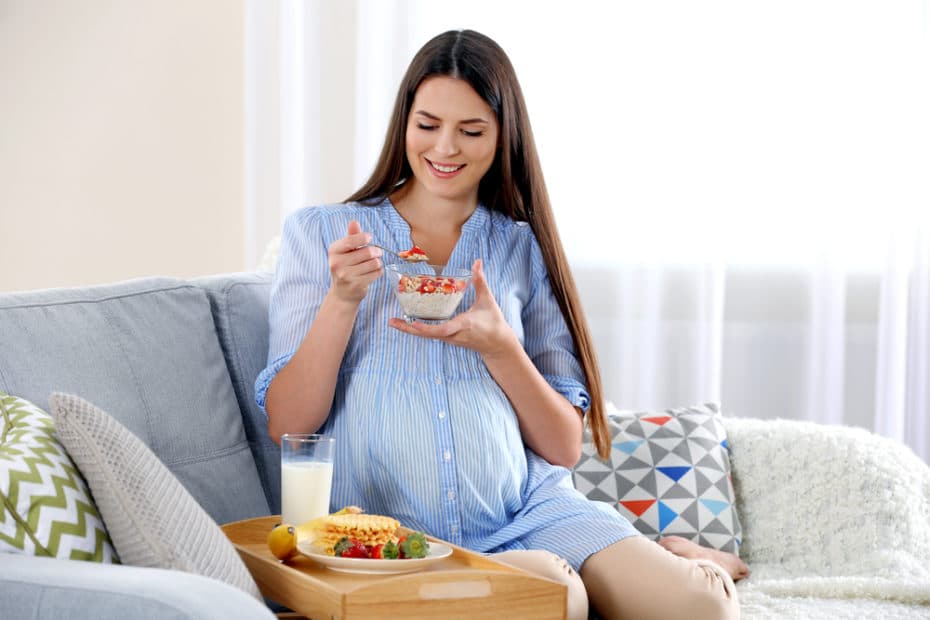 Alimentação na gravidez. Mulher grávida comendo sentada no sofá