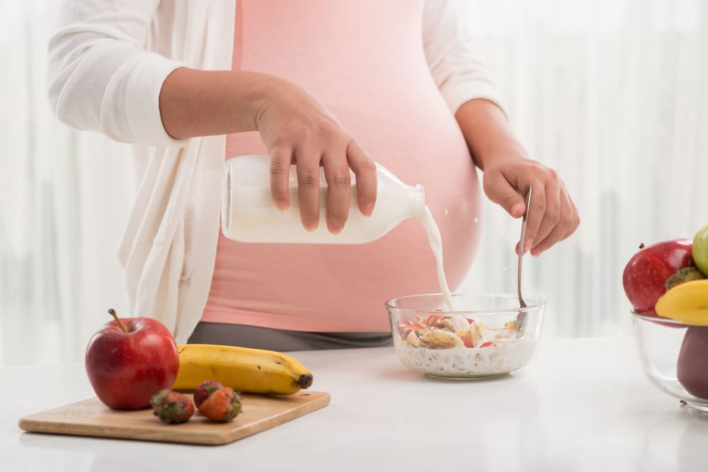 Qual a melhor vitamina para gestante. Mulher grávida colocando leite em uma tigela com cereais e frutas.